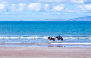 A wide angle shot of two riders with their horses riding on the beach with the clear ocean water behind them