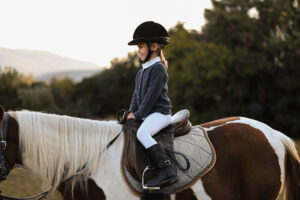 A little girl learning to ride in white jodphurs on a patchy pony with trees in the background